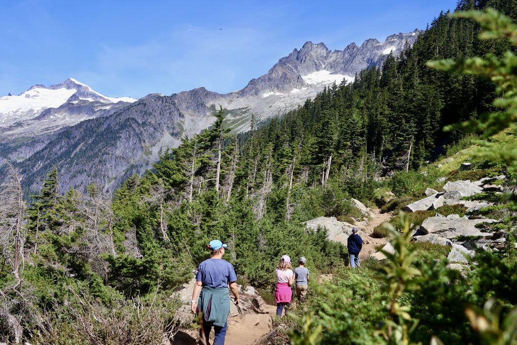 Seen from behind, an adult and three children walk a rocky trail through evergreens toward jagged, snow-patched peaks on the way to Cascade Pass.
